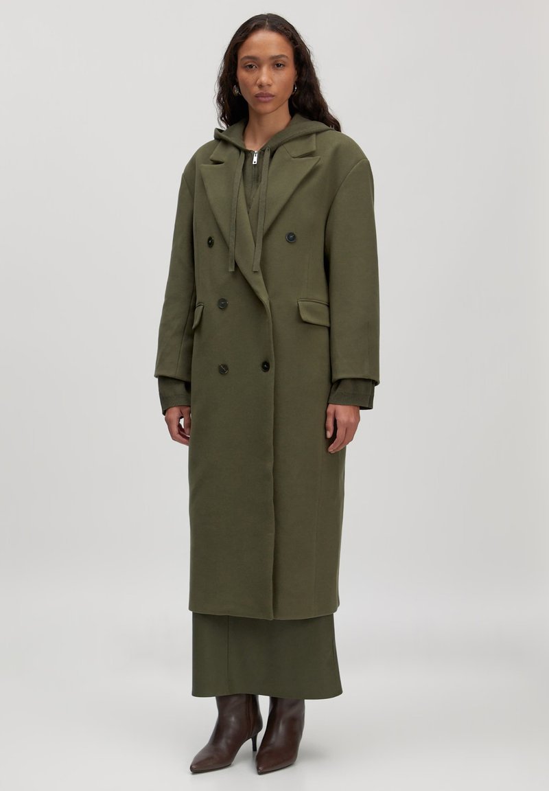 Olivgrüner, doppelt knöpfbarer Mantel mit Kapuze, großen Revers, seitlichen Taschen, kombiniert mit einem passenden Kleid und braunen spitzen Ankle Boots.