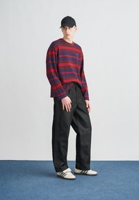 Ριγέ μπλούζα με μακριά μανίκια σε σκούρο μωβ και κόκκινο, συνδυασμένη με μαύρο φαρδύ παντελόνι. Συνδυάζεται με λευκά παπούτσια με σκούρες ρίγες και μαύρο καπέλο.