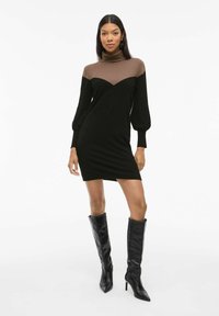 Robe en tricot avec un col roulé marron et un corps noir, présentant un motif en chevrons, de longues manches bouffantes et une silhouette ajustée. Bottes noires à talons hauts.