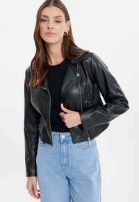 Giacca moto in pelle nera con un design aderente, cerniere sul davanti e ai polsini, colletto con revers, abbinata a jeans azzurri chiari.