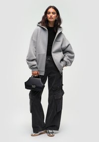 Felpa grigia oversize con tasche anteriori, pantaloni cargo neri, scarpe argentate lucide e una piccola borsa a tracolla nera con un accento metallico.