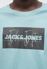 Ανοιχτό μπλε βαμβακερό μπλουζάκι με μαύρο ορθογώνιο λογότυπο που περιέχει λευκό και βεραμάν κείμενο, καθώς και ένα υφασμάτινο φόντο με σχέδιο.