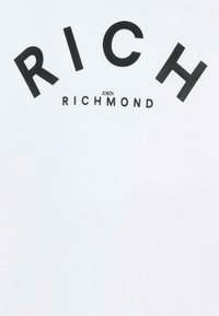 T-shirt bianca con testo nero in grassetto. "RICH" in evidenza, con "JOHN RICHMOND" in un carattere più piccolo sotto. Tessuto morbido.
