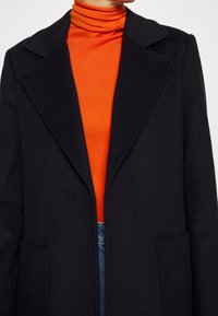 Cappotto di lana nero con rever pronunciato, due tasche frontali, indossato sopra un dolcevita arancione aderente, che mette in risalto i colori a contrasto e il design elegante.