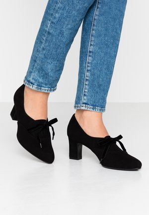Chaussures à talons hauts en daim noir avec un bout pointu, ornées d'un nœud sur le devant, portées avec un pantalon en denim bleu clair.