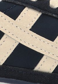 Tejido texturizado en color azul marino oscuro con tirantes cruzados en beige y costuras visibles. Presenta una parte superior suave y una suela de goma. Vista en primer plano.