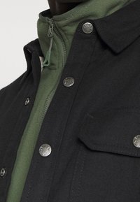 Schwarz geknöpfter Jacke mit silbernen Druckknöpfen, über einem grünen Fleecepullover geschichtet, mit einem Reißverschluss und zwei Brusttaschen.