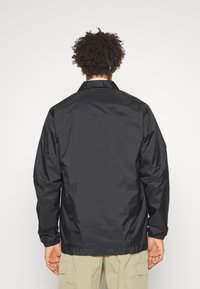 Černá nylonová bunda s límcem, elastickými manžetami a volným střihem. Zadní pohled ukazuje hladkou strukturu a minimalistický design, spojený s lehkými kalhotami.