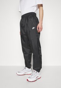 Černé běžecké kalhoty Nike s elastickým pasem, bočními kapsami na zip a logem na levé noze; hladký, lehký materiál.