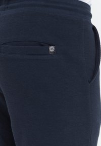 Marineblå joggers med tekstureret stof, der har en sidelomme og et lille cirkulært logo-patch nær taljen.