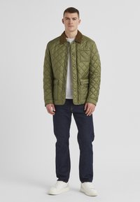 Gesteppten olivgrünen Mantel mit einem braunen Cordkragen, Fronttaschen, Druckknopfverschluss und einem leichten Design, kombiniert mit dunklen Jeans.