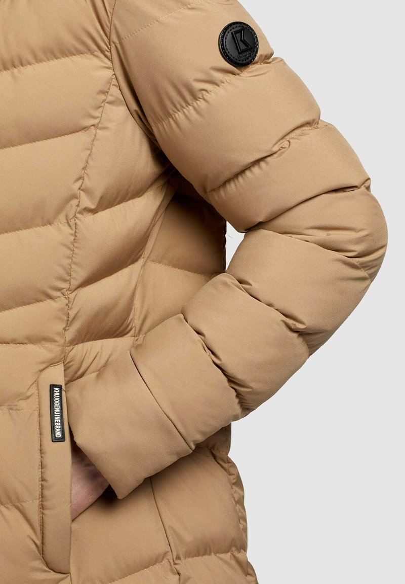 Beige Pufferjacke mit gestepptem Muster, ausgestattet mit einem schwarzen Markenpatch am Ärmel und einer Seitentasche, gefertigt aus glattem, isolierendem Material.