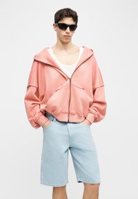 Νεαρός άνδρας που φοράει φαρδιά ροζ φούτερ με φερμουάρ, λευκή αμάνικη μπλούζα, ανοιχτόχρωμα μπλε τζιν σορτς και μαύρα γυαλιά ηλίου, με τα χέρια στις τσέπες.