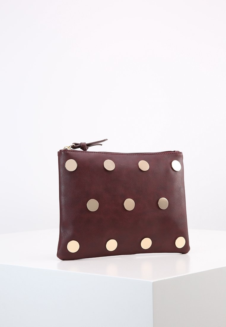 Pochette en cuir marron foncé avec des clous ronds dorés disposés en trois rangées, posée sur un socle blanc devant un fond blanc uni.