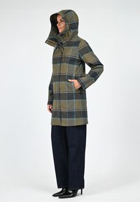 Manteau de pluie long à capuche en vert olive avec un motif écossais noir et bleu ; il possède une fermeture éclair, des poches et est porté sur un pantalon large sombre.