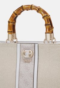 Sac à main beige Guess avec surface texturée, garniture verticale blanche, logo doré et poignées courbées style bambou.