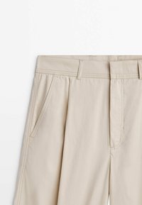 Pantalon beige avec passants de ceinture, poches avant inclinées et coutures visibles le long de la ceinture et des coutures latérales.