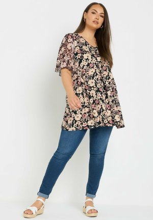 Donna con capelli lunghi castani che indossa una tunica nera con motivi floreali e maniche trasparenti, jeans skinny blu e sandali con zeppa bianchi, in piedi davanti a uno sfondo bianco.