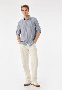 Blauw-wit gestreepte overhemd met opgerolde mouwen, gecombineerd met crèmekleurige broek en lichte sneakers. Soepele stoftextuur.