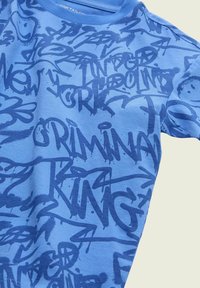 Zila kokvilnas T-krekls ar grafiti stila dizainu, kam raksturīga tumši zila teksts un raksti, tostarp vārdi "Krimins" un "Kungs".