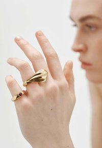 Anillos metálicos dorados en una mano: uno con un diseño liso y curvado, el otro con un acento de gema. Textura suave, formas elegantes, color cálido.