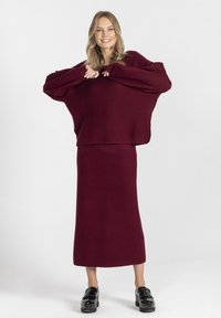 Burgundowy sweter z prążkowanego dzianiny i pasująca spódnica midi z szerokimi rękawami, w komplecie z czarnymi butami na platformie, na białym tle.