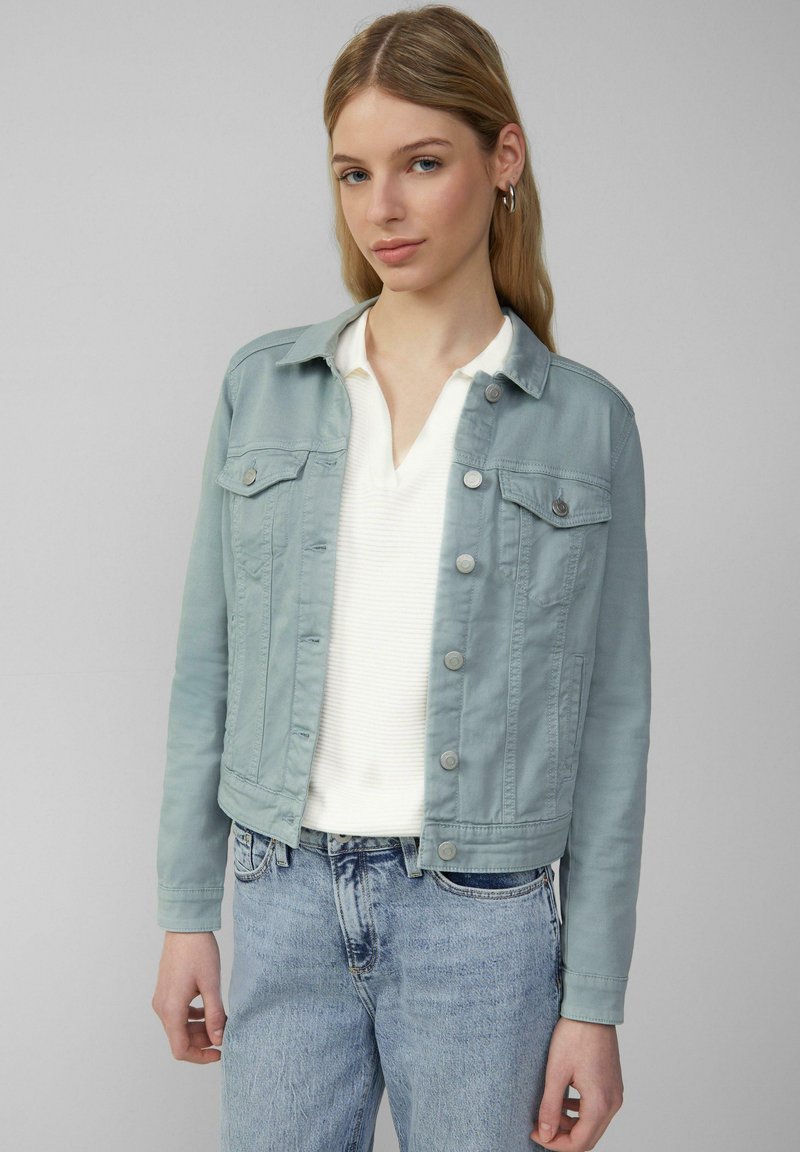 Lichtblauwe denimjack met een knoopsluiting aan de voorkant, borstzakken en een relaxed fit, gedragen over een witte textuurtop en lichtblauwe jeans.