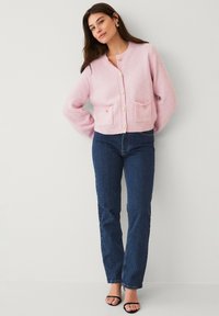 Roze wollen cardigan met een ronde halslijn, knopen aan de voorkant en twee opgestikte zakken. Slank-fitting donkerblauwe jeans maken de outfit compleet.