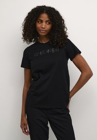 T-shirt en coton noir avec le mot "CHERISH" en sequins noirs brillants sur la poitrine. Manches courtes et coupe décontractée.
