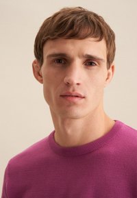 Pullover a maglia ristrutturato di un vivace color viola, con un collo rotondo a costine. Modella con espressione neutra e capelli corti, castano chiaro.