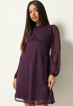 Donna con capelli lunghi e scuri che indossa un vestito viola a maniche lunghe con maniche trasparenti, in piedi davanti a uno sfondo chiaro e semplice.