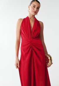 Červené šaty s halter strihom a hlbokým V-výstrihom, skladaným korzetom a voľnou sukňou. Doplnkom je zlatá clutch s textúrovaným povrchom. Hladká tkanina, elegantný dojem.