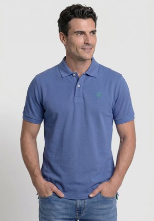 Hombre con una camisa polo azul de manga corta y vaqueros azules, de pie con las manos en los bolsillos, mirando hacia la derecha con una leve sonrisa.