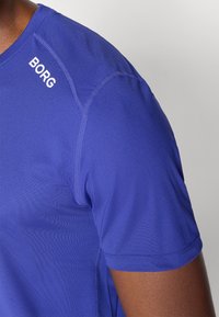 Koszulka sportowa z krótkim rękawem w intensywnym niebieskim kolorze. Posiada okrągły dekolt, gładką teksturę oraz logo "BORG" w białym kolorze na ramieniu.