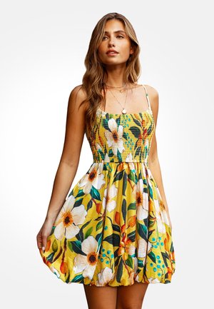 Žena u žutoj haljini bez rukava s velikim bijelim i narančastim cvjetnim uzorcima, stoji nasuprot jednostavnoj pozadini.