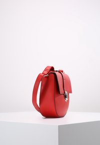 Červená kožená crossbody taška s předním klopovým víkem a stříbrným uzávěrem, stojící vzpřímeně na bílém podstavci na jednolitém pozadí.