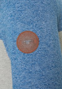 Blauer strukturierter Stoff mit einem melierten Design. Ausgestattet mit einem braunen Lederpatch, der mit "Premium Quality Goods" und einem Warenzeichen-Symbol geprägt ist.