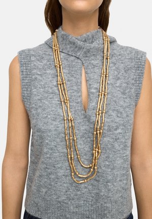 Kobieta w szarym dzianinowym topie bez rękawów z dekoltem typu keyhole i warstwowym, długim naszyjnikiem z złotych koralików.