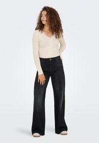 Beige pitkähihaine paita, jossa on pitsireunat ja V-pääntie, yhdistetty korkeavyötäröisiin, leveälahkeisiin mustiin farkkuihin. Malli seisoo yksinkertaista taustaa vasten.