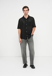 Juoda marškinėlių su trumpomis rankovėmis, priekyje su vertikaliomis juostelėmis ir krūtinės kišene. Derinama su pilkomis plačiomis kelnėmis ir juodais batais.