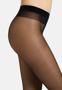 Nahaufnahme einer Person, die durchsichtige schwarze Strumpfhosen mit einem festen schwarzen Bund trägt, sichtbar von der Taille bis zur Mitte des Oberschenkels auf einem schlichten Hintergrund.