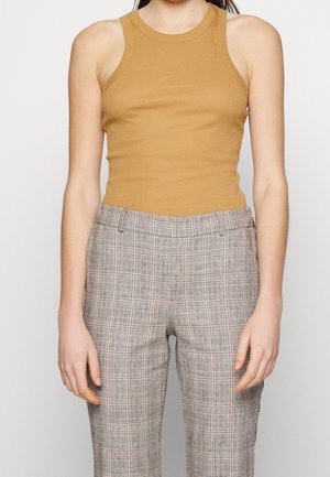 Mujer vestida con una camiseta sin mangas mostaza con textura acanalada y pantalones de cintura alta de cuadros grises, de pie frente a un fondo liso.