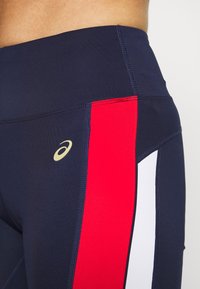 Nærbillede af navy blå sportsleggings med røde og hvide sidelommer samt et lille beige Asics-logo på hoften.
