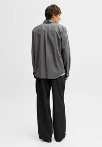 Pelēks garām piedurknēm krekls relaksētā piegriezumā ar aizmugurē esošo jostu detaļu un pogām pie aprocēm, pārī ar brīviem melniem biksēm. Gluda auduma tekstūra.