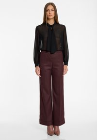 Vrouw in een doorschijnende zwarte blouse met een strik, bordeaux wijde broek met fantasie lijnen, en bruine puntige laarzen, staand op een witte achtergrond.