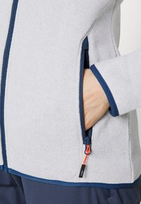 Gray knit jacket with navy accents, featuring a zippered pocket and an adjustable pull tab. Textured fabric and fitted design.