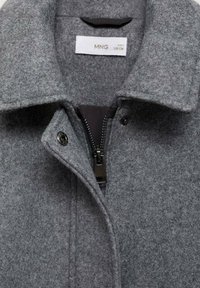 Manteau en mélange de laine gris présentant un col cranté, une fermeture éclair noire, une fermeture à pression et une texture lisse avec une étiquette indiquant la taille 8.