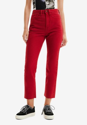 Henkilö, joka käyttää korkeavyötäröisiä lyhytlahkeisia punaisia farkkuja, mustavalkoisia tennareita ja mustaa hihapotonta toppia, jossa on graafinen kuvio.