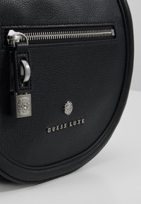 Černá kožená kulatá kabelka s texturovaným povrchem, stříbrným zipem a označením "GUESS LUXE" v kovových písmenech. Obsahuje dekorativní zámek.