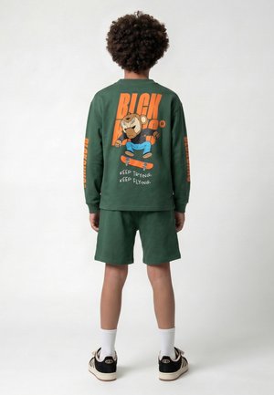 Kind met krullend haar draagt een donkergroen shirt met lange mouwen en korte broek met een skateboardende beer en de tekst "Keep trying, keep flying" op de rug.